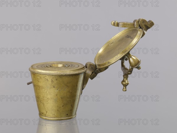 Set of weights. Seven weights in the form of bowls – truncated cones; the eighth, largest, serves as a container for the others and has a hinged lid closed with a hasp and a movable handle mounted in pinnacles shaped like simplified vases. On the lid: 16 MARK, Prussian eagle, hallmark BERLIN; another mark engraved 160; 152 engraved on the side weight 1865.57 g. 18th century, Germany. Weight; height 17.5 cm, width 10.2 cm, depth 14.3 cm., A small metallic object made of brass with a dull golden color is shown against a light gray background; it consists of a tapered cylindrical cup with a concentric ridged lid, a hinged circular plate opened upward, and an attached decorative swing arm ending in a turned finial, all showing aged surface wear and small dark spots, with a short metal pin protruding from the side of the cup.