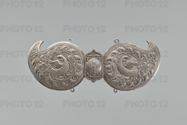 Buckle, Two-part silver buckle fastening Delya's coat, probably royal; inscription Warszawa / 24 11 1705 engraved in italics on one vertical strip on the reverse. Each element shaped like a bota with a repoussé border of pearl-strips and leaves; inside, circular medallions with a phoenix holding snakes in its beak. The elements are connected with a hook; the part with the ring is covered with a rectangular shield with ogee spines top and bottom, bearing a circular medallion with the Polish eagle. Both parts have soldered rings at top and bottom; vertical strips for attaching a belt are soldered on the reverse; one has a disc on the clasp. Not signed, not hallmarked. Possibly a Middle Eastern work from the 19th century, later supplemented with a disc with the Polish eagle and the inscription, probably made in Europe in the third quarter of the 19th century. Germany place. Clothing accessory — buckle. Height 10.5 cm, width 26.5 cm., A decorative metallic object with a pair of symmetrical...