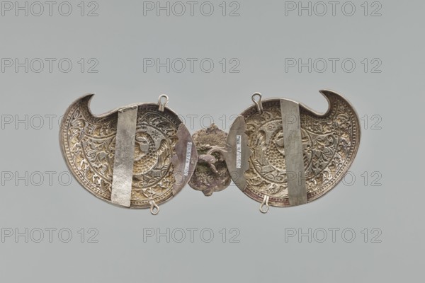 Buckle, Repossession; gift. Silver buckle fastening Delya's coat, probably royal, inscription 24 February 1705. Two-part, each element in the form of a bota, with a repoussé border composed of strips of pearls and leaves. Inside, circular medallions with a phoenix holding snakes in its beak. Both elements are connected with a hook, the part with the ring covered with a rectangular shield with ogee spines at top and bottom, bearing a circular medallion with the Polish eagle. Both parts at top and bottom have soldered rings; on the reverse, vertical strips for attaching a belt are soldered on; one of them with a disc on the clasp is engraved in italics with the words Warszawa / 24 11 1705. Not signed, not hallmarked. The buckle may be a Middle Eastern work from the 19th century, later supplemented with a disc with the Polish eagle and an inscription, probably from the third quarter and made in Europe. 3rd quarter of the 19th century; clothing accessory buckle; height 10.5 cm, width 26.5 ...