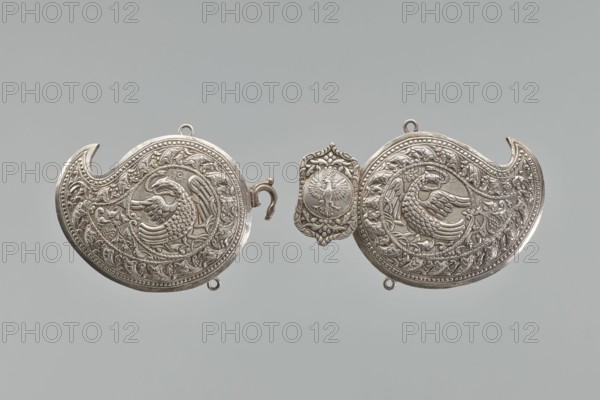 Buckle, repossession, gift. Silver buckle fastening Delya's coat, probably royal, inscription 24 February 1705. Two-part buckle, each element in the form of a bota, with a repoussé border composed of strips of pearls and leaves. Inside, circular medallions with a phoenix holding snakes in its beak. Both elements are connected with a hook; the part with the ring is covered with a rectangular shield, with ogee spines at the top and bottom, bearing a circular medallion with the Polish eagle. Both parts at the top and bottom have soldered rings; on the reverse, vertical strips for attaching a belt are soldered on; one of them with a disc on the clasp is engraved in italics with the words Warszawa / 24 11 1705. Not signed, not hallmarked. The buckle may be a Middle Eastern work from the 19th century, later supplemented with a disc with the Polish eagle and an inscription, probably from the third quarter and made in Europe. 3rd quarter of the 19th century; Germany. Clothing accessory buckle....