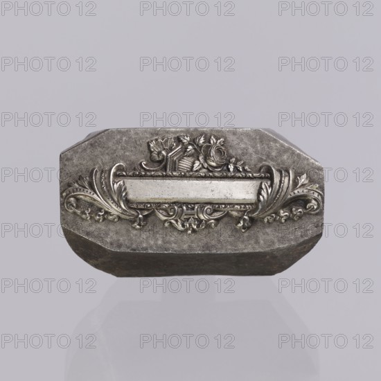 Jewellery die, Approximately hexagonal die with a relief rectangular reserve enclosed at the ends by two leaves. At the top and bottom of the reserve are floral and plant decorations and scrolls. Probably a jeweller's die for making semi-finished products, 2nd half of the 19th century, Germany. Die; height 3 cm, width 6 cm, depth 3 cm., A metallic, octagonal object sits against a pale gray background, showing a raised, ornate central plaque with a horizontal blank panel flanked by symmetrical scrollwork and leafy flourishes; above the panel is a decorative motif including a rose and ribbon-like elements, and the metal surface displays a textured, slightly worn finish with darker areas along the lower edge.