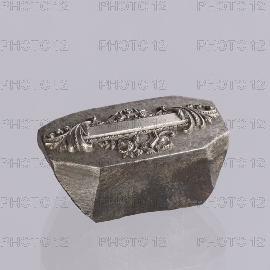 Jewellery die, Approximately hexagonal die with a relief rectangular reserve enclosed at the ends by two leaves. At the top and bottom of the reserve are floral and plant decorations and scrolls. Probably a jeweller's die for making semi-finished products, 2nd half of the 19th century, Germany. Die; height 3 cm, width 6 cm, depth 3 cm., A single irregularly shaped metallic object sits against a pale gray background, showing a textured, dark gray base with areas of rust-colored brown and lighter gray wear. The top surface features a raised decorative band with ornate scrollwork and floral motifs in a shinier silver-gray, exhibiting highlights and small shadows that emphasize its carved details. The object casts a soft reflection and faint shadow on the surface beneath it.