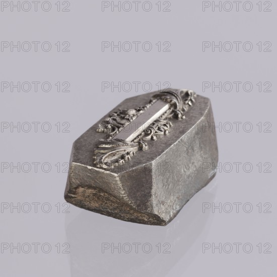 Jewellery die, Approximately hexagonal die with a relief rectangular reserve enclosed at the ends by two leaves. At the top and bottom of the reserve are floral and plant decorations and scrolls. Probably a jeweller's die for making semi-finished products, 2nd half of the 19th century, Germany. Die; height 3 cm, width 6 cm, depth 3 cm., A roughly rectangular metallic object with faceted sides sits on a smooth surface, featuring an ornate raised decorative element on its top with carved scrollwork and a central polished strip; the object shows a mix of matte and slightly reflective textures with visible wear and rough edges, and the visible colors are shades of gray, silver, and a bit of muted brown on one lower edge, set against a pale lavender-gray background.