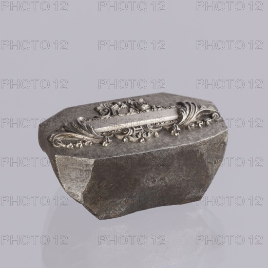 Jewellery die, Approximately hexagonal die with a relief rectangular reserve enclosed at the ends by two leaves. At the top and bottom of the reserve are floral and plant decorations and scrolls. Probably a jeweller's die for making semi-finished products. 2nd half of the 19th century, Germany. Die; height 3 cm, width 6 cm, depth 3 cm., A roughly rectangular dark gray stone with uneven, chipped edges sits against a light gray background, showing a decorative silver-colored metal applique on its top face that features ornate scrollwork, floral motifs, and curved leaf-like forms; the stone surface displays textured gray and brown areas with lighter gray worn patches, while the metal decoration has a polished silvery sheen with small shadowed recesses.