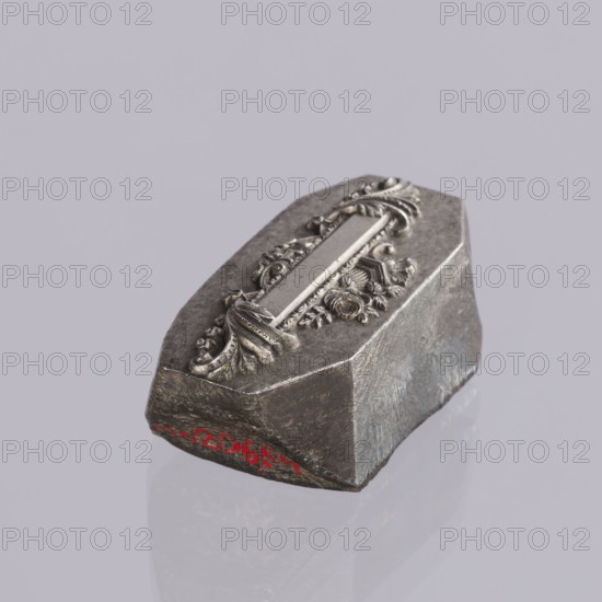 Jewellery die, Approximately hexagonal die with a relief rectangular reserve enclosed at the ends by two leaves. At the top and bottom of the reserve are floral and plant decorations and scrolls. Probably a jeweller's die for making semi-finished products, 2nd half of the 19th century, Germany. Die; height 3 cm, width 6 cm, depth 3 cm., A small metallic object rests on a smooth reflective surface, showing a faceted base with rough textured sides and a top adorned with an ornate, raised decorative element featuring curved leaf-like and floral motifs; the colors visible include shades of gray, silver, dark metallic tones, and a small area of red along one lower edge, set against a pale gray background.