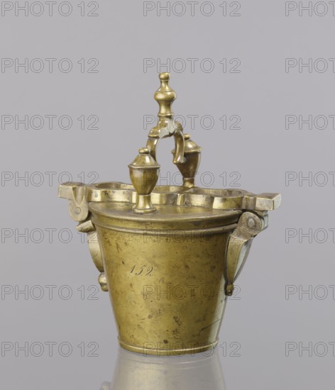 Seven weights in the form of bowls—truncated cones; the eighth largest serves as a container for the others and has a hinged lid. Closed with a lid, with a movable handle mounted in pinnacles shaped like simplified vases. On the lid stamped 16 MARK, Prussian eagle, BERLIN stamp; 160 engraved on another mark; 152 engraved on the side. Late 18th century, Germany. Weight. Creator: Gwicht, Szewczykowski, Roman 1849–1901., A small metal bucket-like vessel with a hinged decorative lid and handle, shown against a plain light gray background; the object is metallic brass in color with areas of darker tarnish and patina, featuring rounded finials and curved ornamental supports, a rope-like rim detail, two small projecting bracket-like elements on the sides, and a reflective surface beneath that shows a faint mirrored outline.