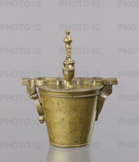 Seven weights in the form of bowls—truncated cones; the eighth, largest, serves as a container for the others and has a hinged lid with a movable handle mounted in pinnacles shaped like simplified vases. On the lid: stamped 16 MARK, Prussian eagle, BERLIN stamp; another mark engraved 160; on the side engraved 152. Late 18th century, Germany. Materials: measures and weights. Maker: unknown., A brass container with a domed lid and a turned finial on top, showing a warm golden color with areas of darker patina and small spots. The container has a tapered cylindrical body and a decorative rim beneath the lid with a subtle rope-like pattern, and a pair of hinged bracket-like handles on either side with rounded end pieces, all in the same brass tones. The lid has an irregularly shaped flat top section with cutouts around the central finial, and the finial itself is a turned, vase-like form stacked with rounded and cylindrical segments. The background is a uniform light gray and the object re...