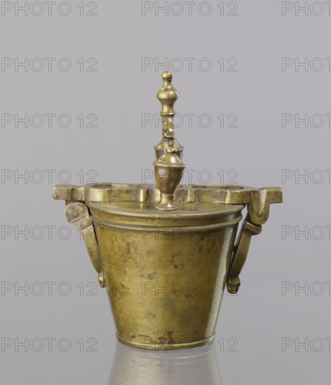 Seven weights in the form of bowls — truncated cones; the eighth largest serves as a container for the others, with a hinged lid. Closed with a lid, with a movable handle mounted in pinnacles shaped like simplified vases. On the lid stamped 16 MARK, Prussian eagle, BERLIN stamp; 160 engraved on another mark; on the side engraved 152. Late 18th century, Europe, Germany. Weight., A brass-colored metal vessel with a conical body and a round base sits against a pale gray background, showing a slightly worn and mottled surface with darker spots and patina. The vessel has a decorative lid topped by a finial composed of stacked rounded shapes and a small knob, and a horizontal rim with a ridged band where the lid meets the body. Hinged bracket-like extensions project from the rim on opposite sides, each with articulated arms attached near the upper body. Reflections of the object appear faintly on the smooth surface beneath it.