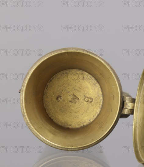 Seven weights in the form of bowls — truncated cones; the eighth, largest, serves as a container for the others and has a hinged lid with a movable handle mounted in pinnacles shaped like simplified vases. On the lid: stamped 16 MARK, Prussian eagle, BERLIN stamp; another mark engraved 160; on the side engraved 152. Late 18th century, Germany. Weight. Creator: Gwicht, unknown., A close-up view of the inside of a round metal cup with a hinged lid visible at the right edge; the interior shows a worn, textured surface in shades of gold and brass with darker tarnished spots and a few rough patches, while the exterior rim and hinge also display metallic gold and brass tones, and the background is a smooth pale gray.