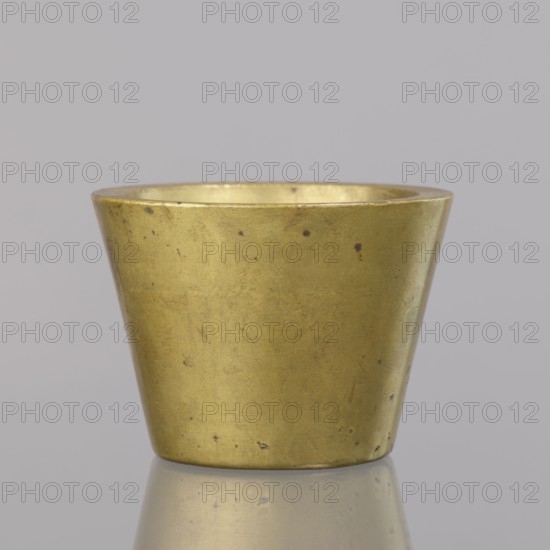 Gwicht, Szewczykowski, Roman 1849–1901 — measures and weights; in the shape of a truncated cone; markings: 4 M engraved inside and on the side 76; weight 935.18 g; end of 18th century; Germany; height 6.2 cm; width 8.6 cm, A small metallic cup with a tapered cylindrical shape sits centered against a plain background; the cup is a muted gold color with a slightly worn and tarnished surface showing a few dark spots and small scratches, and its inner rim is visible with the same gold tone; the cup rests on a reflective surface that mirrors its base and shows faint muted gold and gray reflections; the background is a pale grayish color.