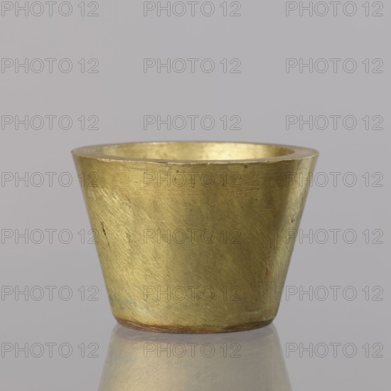 Gwicht; creator, Szewczykowski, Roman 1849–1901 — . Measures and weights: in the shape of a truncated cone, without markings; bottom surface covered with a layer of tin; weight 410.2 g; end of the 18th century; weight; height 4.9 cm; width 6.9 cm., A small metallic cup with a tapered cylindrical shape in a golden brass color, showing a slightly worn and scratched surface with subtle darker spots and patina; the rim is visible and the interior appears the same golden color; the base edge has a thin band of brownish discoloration and the cup sits on a reflective surface that shows a muted reflection; the background is a smooth light gray, and overall visible colors include gold, brown, gray, and hints of darker greenish patina.