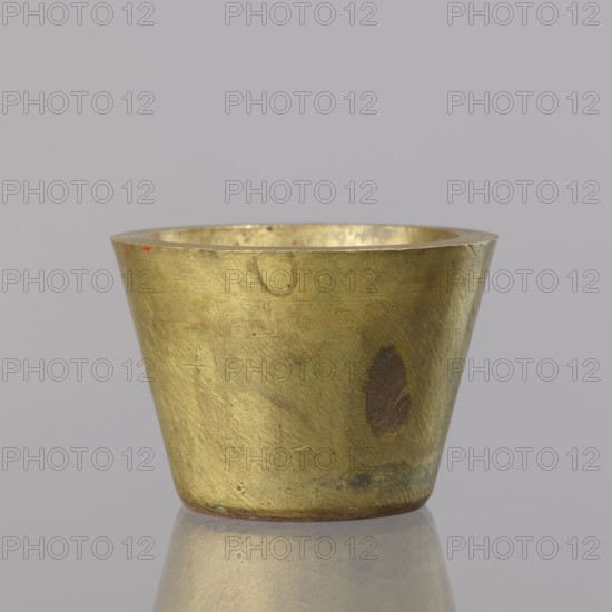 Gwicht; creator, Szewczykowski, Roman 1849–1901 — ; measures and weights; object in the shape of a truncated cone; without markings; bottom surface covered with a layer of tin; weight 410.2 g; late 18th century; Berlin; height 4.9 cm; width 6.9 cm, A small metallic cup with a tapered cylindrical shape sits centered on a reflective surface, showing a worn golden brass color with areas of darker brown tarnish and a few small spots of greenish corrosion, the metal surface displaying fine scratches and scuffs, and a faint reddish mark near the rim; the background is a uniform pale gray.