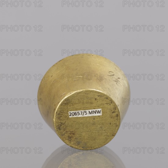 Gwicht;, In the shape of a truncated cone; engraved marking on the side 91/2, inside 8, eagle; 116.45 g; end of 18th century; Europe, Germany; weight; height 2.9 cm; width 4.5 cm., A cylindrical metal object viewed from one circular end, showing a worn, slightly rough surface with scratches and small dark spots; the metal has a yellowish-gold color with areas of darker discoloration and some lighter highlights; it rests on a smooth, reflective gray surface that creates a faint reflection below; the background is a uniform pale gray.