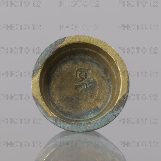 Gwicht, unknown, Szewczykowski, Roman 1849-1901 — In the shape of a truncated cone, internal marking 2, weight 24.75 g, 18th century, Europe, Germany, height 1 cm, width 3.1 cm, A round metal object with a shallow recessed interior, showing worn surfaces and an indented decorative mark in the center; the rim is thick and uneven with areas of wear and patina. The visible colors include brassy gold, dark brown, greenish-blue verdigris, and muted gray tones. The object sits against a light gray background and its underside is reflected faintly on a glossy surface below.