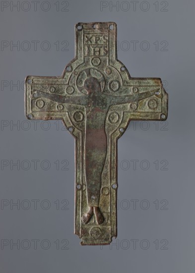 Cross from the binding of a liturgical book or a processional, Romanesque, beginning of the 13th century, possibly from Limoges, France. Enamelled originally now without traces of enamel. Relief image of Christ with the inscription XPS/IHS; only the feet of the Crucified figure remain. Described as either a plaque from a book binding or a processional cross the latter likely. Possibly originated from Kacice, connected with Cistercians from Mogila. Cross, height 21 cm, width 13.5 cm., An aged metal cross-shaped plaque with a central figure in low relief; the figure's head, torso, and legs are visible, with the feet positioned together at the bottom. The plaque shows a ringed circular motif around the figure's head and several circular raised decorations across the arms and body area. There are small round holes near the edges and along the vertical axis. The surface exhibits a weathered, mottled patina with predominant green and brown tones, with hints of darker reddish-brown on the fig...