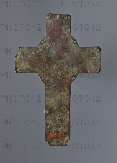 Cross from the binding of a liturgical book or processional, liturgies, processions, enamelling, Romanesque cross with a relief image of Christ and the inscription XPS/IHS. Originally enameled, currently without traces of enamel. Only the feet of the Crucified figure remain. Found in 1911 in a field near Slomniki then within the Kingdom of Poland. Described as a plaque from the binding of a liturgical book; possibly a processional cross. May come from Kacice, which belonged to the Cistercians who later settled in Mogila. Beginning of the 13th century, Limoges, France. Cross, height 21 cm, width 13.5 cm., A weathered metal cross-shaped plate with irregular edges and a group of small circular holes near the ends and along the body, showing a mottled surface with patches of green, brown, dark reddish-brown, and hints of yellowish discoloration, set against a plain neutral gray background.
