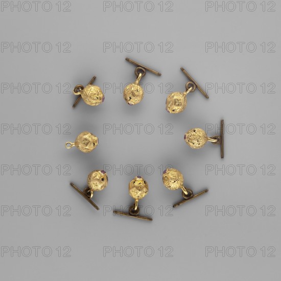 Buttons for a zupan with clasps. Nearly spherical, polygonal buttons with engraved four-leaf rosettes inside. Each features a table-cut ruby in the chest; in the surrounding fields, a stylized acanthus leaf, enameled in black. On the opposite side, oval lugs are soldered on, some with secondary clasps – brass drums. The dating element is the enameled decoration; the form and this type of engraved decoration were repeated for a long time. Baroque style; rosette, acanthus, geometric ornament. 17th–18th century, Poland, clothing accessories, weight 15.95 g., A circular arrangement of small spherical charms with textured faceted surfaces in a metallic gold color, each attached to short cylindrical bars and rings in a darker bronze tone, set against a smooth neutral gray background; a few of the gold spheres show tiny accents of deep red or purple on one side.