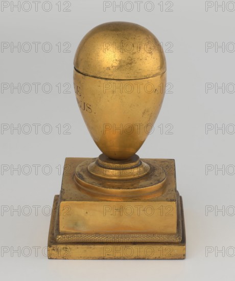 Inkwell, maker, Tadeusz Bylewski 1866–1939 personal entry; early 19th century, France, inkwell; height 13.5 cm, width 9.4 cm, depth 8.4 cm, A golden egg-shaped object with a rounded top sits on a stepped circular pedestal atop a square base, showing a worn and slightly mottled surface with areas of darker patina and small scratches; the base has a textured border and flat panels, and the overall colors visible are gold with darker brownish spots and hints of muted gray in the background.
