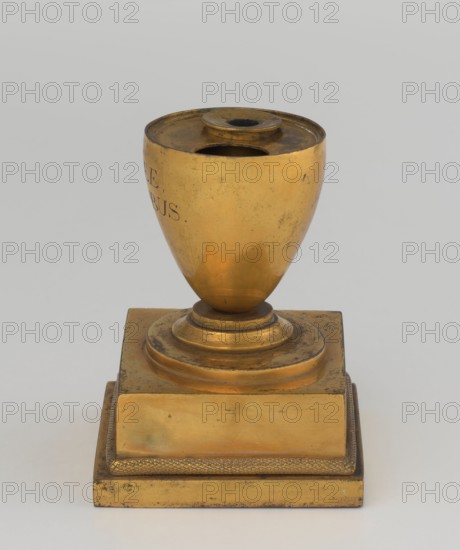 Inkwell, early 19th century, France, inkwell, height 13.5 cm, width 9.4 cm, depth 8.4 cm, A metallic object with a golden color sits on a flat surface against a neutral gray background, consisting of an urn-shaped upper section with a round recessed top and a central circular opening, mounted on a stepped square base that also appears golden and shows areas of darker tarnish and small spots of wear, with a textured band around the lower tier and subtle reflections and shadows on the smooth curved surfaces.