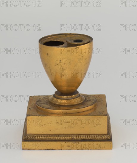 Inkwell, early 19th century, France, inkwell; height 13.5 cm, width 9.4 cm, depth 8.4 cm., A gilt metallic object with a rounded cup-like top featuring two open circular cavities sits on a decorated stepped base; the object shows worn and scratched surfaces with patches of darker tarnish and a few areas of black marks. The visible colors are gold, darker brownish tarnish, black, and a pale gray background.