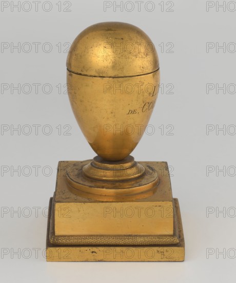 Inkwell, early 19th century, France, by Tadeusz Bylewski 1866–1939, inkwell; height 13.5 cm, width 9.4 cm, depth 8.4 cm, A gilded object resembling an egg-shaped vessel with a rounded top sits on a stepped square base; the vessel has a smooth, slightly weathered metallic surface with areas of darker tarnish and small scratches, and the base shows similar wear with a textured border detail. The visible colors are gold and darker golden-brown tones against a pale gray background.