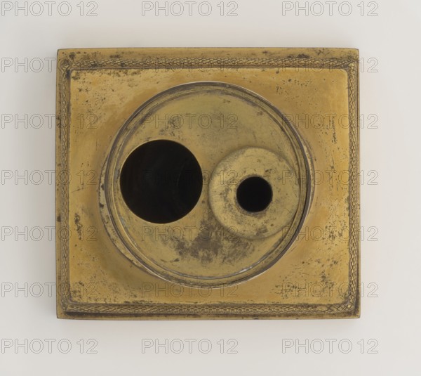 Inkwell, early 19th century, France, inkwell, height 13.5 cm, width 9.4 cm, depth 8.4 cm, A square metal object with a raised decorative border and a central circular recessed area containing two round openings, one larger and one smaller; the surface shows tarnish and wear with mottled patches. The visible colors are gold, dark brown, black, and hints of gray.