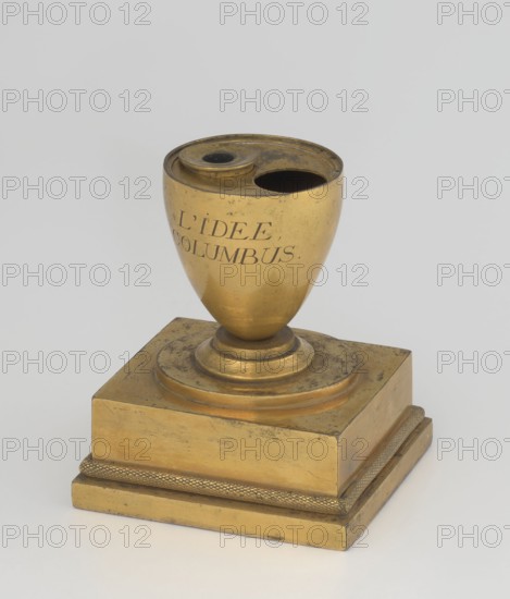 Inkwell, early 19th century, France, inkwell, height 13.5 cm, width 9.4 cm, depth 8.4 cm, A golden brass object shaped like an egg cup mounted on a square stepped base, with a round recessed top that has two openings and a small raised rim; the surface shows areas of darker tarnish and patina alongside brighter gold highlights, and the base includes a textured band around its lower edge.