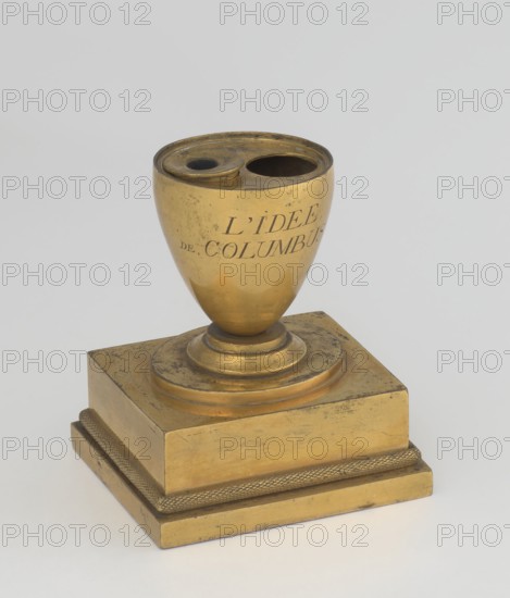 Inkwell, early 19th century, France, maker, attributed to Tadeusz Bylewski 1866–1939; inkwell; height 13.5 cm, width 9.4 cm, depth 8.4 cm., A small metallic object sits on a flat surface, composed of a rounded cup-like top mounted on a stepped square base. The cup portion has two round openings at its rim and shows a smooth, reflective surface with some scuffs and dark spots. The base is tiered with a textured band around its lower tier and displays worn areas and surface marks. The overall color palette is primarily gold with darker tarnished areas and small blackish spots, set against a pale gray background.