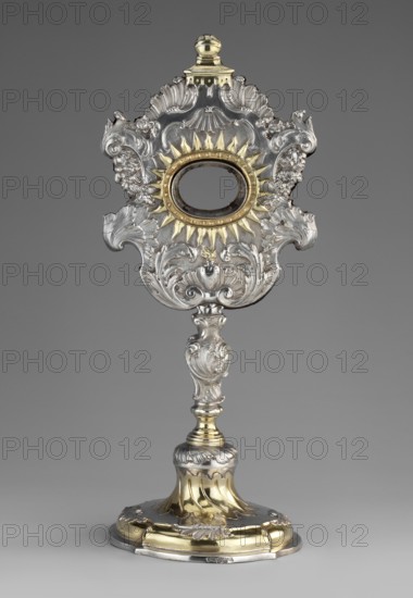 Monstrance reliquary by Johann Gottfried Schlaubitz fl. 1733–1771, goldsmith; originally containing relics of the Holy Cross. Rococo silver reliquary in the shape of a monstrance, supported on a circular, quadripartite, vertically ribbed profiled foot decorated with rooster combs, passing into a shaft with a pear-shaped rocaille nodus; a glory decorated with rocailles and garlands; center oval cutout for relics surrounded by rays, below it a flaming heart engraved with the letters IRS and the Zejbersdorf von Sartawski coat of arms. Inscribed on the foot: Pro Ecclesia Obizinenzi 1748 ca. 1748. Materials: silver; style: rocaille/Rococo. Subject/iconography: Entombment iconography, relics of the Holy Cross; function: reliquary/monstrance. Place of origin: Gdansk, Royal Prussia, Poland. Dimensions: height 31 cm, width 13.5 cm, depth 12 cm., An ornate metallic object stands against a plain gray background, featuring a decorative top section with a central oval opening surrounded by radiatin...