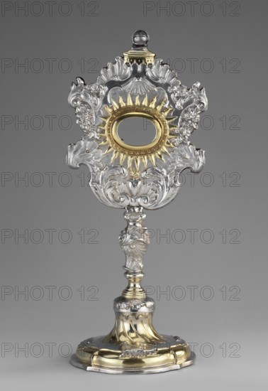 Monstrance reliquary by Johann Gottfried Schlaubitz fl. 1733–1771, goldsmith; originally containing relics of the Holy Cross; rococo silver reliquary in the shape of a monstrance, supported on a circular quadripartite profiled foot with vertical ribs decorated with rooster combs, passing into a shaft with a pear-shaped rocaille nodus; a glory decorated with rocailles and garlands; center oval cutout for relics surrounded by rays, below it a flaming heart engraved with the letters IRS and the Zejbersdorf von Sartawski coat of arms. Inscription on the foot: Pro Ecclesia Obizinenzi 1748 ca. 1748. Provenance: church in Obozin Obozin; Starogard County. Materials: silver; function: reliquary. Dimensions: height 31 cm, width 13.5 cm, depth 12 cm., A decorative metallic object stands upright against a plain gray background, featuring an ornate top section with elaborate scrollwork and sculpted floral and shell-like motifs in silver and gold tones, a central oval opening encircled by a sunburst...
