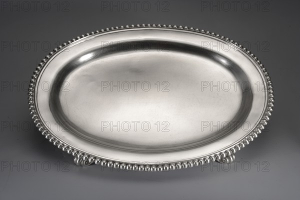 Oval tray by Jacob Haase 1704–1762, goldsmith active from 1730; died 1762; widow active to 1774. Smooth oval tray with a raised collar bordered with pearling, supported by four acanthus legs feet not original, soldered on in the 19th century. Scrap — purchase; purchase provenance. Ornament: pearling and acanthus leaves. Dated 1750, Gdansk, Royal Prussia, Poland. Vessel. Height 3.7 cm; width 35 cm; depth 27.2 cm; weight 1035 g., A large oval metal tray with a smooth reflective surface and a raised rim decorated with a beaded edge rests on a flat surface, showing a few small ornamental feet beneath the rim; visible colors include silver tones on the tray and a muted gray background.