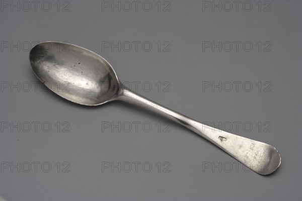 Table spoon with shooting brotherhood emblem, Hermann, Christoph fl. ca. 1740–1778, goldsmith, active from the 1740s, died 1778. Silver spoon, ca. mid-18th century. On the reverse of the bowl: dotted italic inscription Anno 1766 1 April above an engraved shooting shield, crossed rifles, and the Elblag coat of arms. Along the handle: dotted German inscription, very faded in italics Hier isst ? der Platz da zeigt deinen Fleis / Ein Edlen Rath # dir Ehr und Preis. On the obverse of the handle: later inscription AW CH / 1860 / 14 Sept. Function: shooting award presented at annual marksman fraternity competitions. Place: Elblag, Royal Prussia. Type: cutlery. Length 22.5 cm; weight 64 g., A metal spoon lies diagonally across a flat surface, showing a reflective, slightly tarnished bowl and a long handle that tapers to a rounded end; the spoon surface has small spots and scratches and a subtle sheen, and the background and overall scene include shades of gray with areas of lighter and darker ...