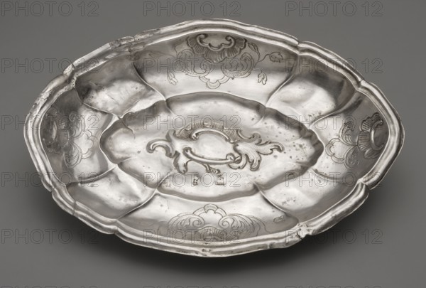 Oval cake stand, maker: Johann Constantin Lange 1702–1761; silver; rocaille and acanthus ornamentation, rooster's comb motifs. Oval silver cake stand with multi-leaf cut, set on four legs in the form of palmettes; a concave-convex mirror with a multi-leaf outline and rocaille decoration, a wavy collar vertically divided by ribs rising upwards, decorated on the longer and shorter axes with four rooster combs and an acanthus twig, and a stepped-profile rim with a multi-leaf outline. Trays of this type, popular in Gdansk goldsmithing of the period, may have been used for serving spoons. Ca. 1763; Gdansk, Royal Prussia Poland. Vessel; weight 130 g; height 4 cm; width 14.8 cm; depth 21.1 cm., A shallow oval metal dish with a scalloped rim sits against a neutral gray background; the dish has a reflective silver surface with areas of darker tarnish and small spots, a central raised decorative cartouche with flowing scroll-like motifs, and a few engraved floral or shell-like designs around the...