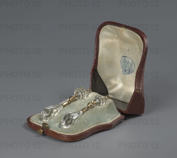 Pair of earrings in a leather case. Made by Sazikov goldsmith's firm; 1793–1887. Pair of earrings made of 56 gold with a faceted rock crystal teardrop set in gold and suspended from a faceted crystal ball. 19th century, St. Petersburg, Russia. Jewelry., A small open case with a dark reddish-brown exterior and a pale cream satin interior holds a soft pale green velvet pad on which a pair of clear faceted teardrop-shaped crystal pendants rest; the pendants are attached to short segments of metallic gold-colored fittings and small round gold-colored beads, and the crystals display multiple flat facets that reflect light. The background is a uniform muted gray.