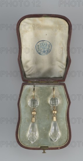Pair of earrings in a leather case. Maker: Sazikov goldsmith's firm, Moscow and St. Petersburg; active 1793–1887. Materials: 56 gold and faceted rock crystal teardrop set in gold, suspended from a faceted crystal ball. Date: 19th century. Place: St. Petersburg, Russia. Function: jewelry., An open hinged case with a brown exterior and pale greenish interior lining holds a pair of dangling earrings; the interior lid is a creamy off-white with some small stains and a blue circular mark, while the lower interior is a soft pale green velvet with slight discoloration. The earrings feature clear faceted round beads at the top, small gold-colored spherical beads below them, tapered gold-colored metal connectors, and elongated clear faceted teardrop pendants at the bottom. The visible colors include brown, creamy off-white, pale green, blue, clear/transparent, and gold.