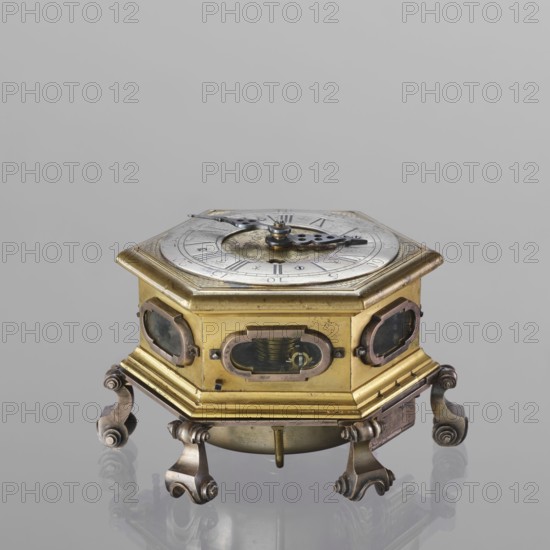 Tiled clock, Michael Gugenmus 1705–1775 watchmaker; rococo, Regency ornament. Table clock, tiled, hexagonal, of gilded bronze on six volute legs, glazed side walls; silver-plated ring-shaped dial with black Roman numerals for hours and Arabic minutes, with two winding holes; center engraved Regency-Rococo cartouche signed Gugenmos/Varssavie; hinged flap closing the mechanism; front plate of the mechanism with a modest bun and a hammer with an engraved bronze head. Signature: Johann / Michael / Gugenmus / A Varsovie. Mechanism with a strike repeater and a handle on the side wall under the upper edge. 2nd half of the 18th century, Warsaw, Poland. Timepiece. Height 8.5 cm; width 13.5 cm; depth 13.5 cm., A hexagonal brass and metal object with a circular dial on top sits on a reflective surface; the top dial is silver-toned with darker metal hands and decorative cutouts, and the hexagonal body shows brass panels with several oval glazed openings revealing internal components. The piece res...