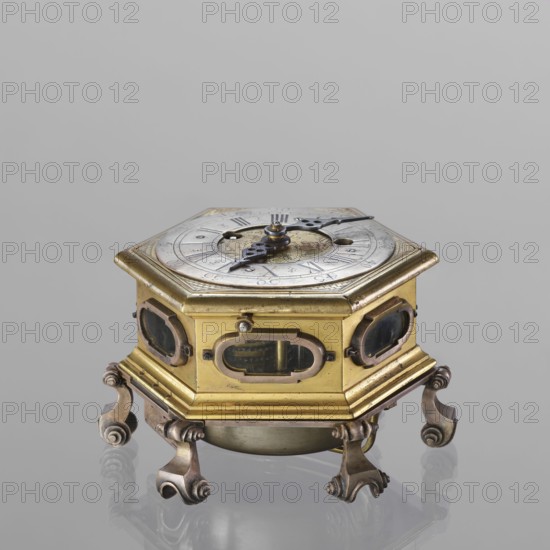Tiled clock, Michael Gugenmus 1705–1775 watchmaker; rococo style with Regency ornament; table tiled clock, hexagonal, gilded bronze on six volute legs, glazed side walls; silver-plated ring-shaped dial with black Roman numerals for hours and Arabic minutes, two winding holes; center engraved Regency-Rococo cartouche with the signature Gugenmos/Varssavie; hinged flap closing the mechanism; front plate of mechanism with a modest bun and a hammer with an engraved bronze head. Signature: Johann / Michael / Gugenmus / A Varsovie. Mechanism with strike repeater, with a handle on the side wall under the upper edge. 2nd half of the 18th century, Warsaw, Poland. Timepiece. Dimensions: height 8.5 cm, width 13.5 cm, depth 13.5 cm., A hexagonal metal object with a metallic top showing a circular dial and two ornate hands sits on a matching hexagonal body with small oval windows in each side; the body is mainly gold with darker metal accents and a few areas of patina, and it rests on a ringed base ...