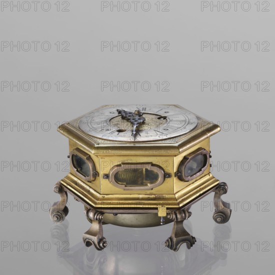 Tiled clock, Michael Gugenmus 1705–1775 watchmaker; rococo style with regency ornament; table tiled clock, hexagonal, gilded bronze on six volute legs, glazed side walls; silver-plated ring-shaped dial with black Roman numerals for hours and Arabic minutes, two winding holes; center engraved Regency-Rococo cartouche with signature Gugenmos/Varssavie; hinged flap closing the mechanism; front plate of mechanism with a modest bun and a hammer with an engraved bronze head; signature: Johann / Michael / Gugenmus / A Varsovie; mechanism with strike repeater and a handle on the side wall under the upper edge; 2nd half of the 18th century; Warsaw, Poland; timepiece; height 8.5 cm, width 13.5 cm, depth 13.5 cm., A small hexagonal brass box with an ornate circular metal dial on top sits on a reflective surface, supported by a group of decorative curved metal feet with scroll details; the top dial has a pierced, dark metal hand and engraved concentric rings, while the box sides feature several sm...