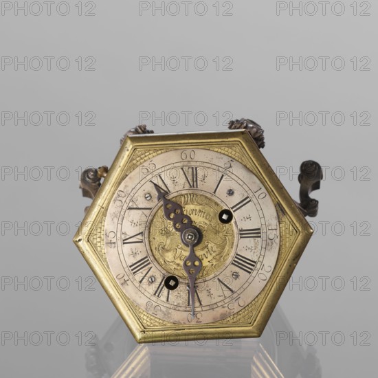Tiled clock by Michael Gugenmus Johann Michael Gugenmus 1705–1775, watchmaker. Rococo, Regency ornament. Hexagonal table clock of gilded bronze on six volute legs with glazed side walls; silver-plated ring-shaped dial with black Roman numerals for hours and Arabic minutes, two winding holes; center engraved Regency-Rococo cartouche signed Gugenmos/Varssavie also given as Johann / Michael / Gugenmus / A Varsovie. Hinged flap closing the mechanism; front plate with a modest bun and a hammer with engraved bronze head. Mechanism with strike repeater and a handle on the side wall under the upper edge. 2nd half of the 18th century. Poland, Warsaw. Timepiece. Height 8.5 cm, width 13.5 cm, depth 13.5 cm., A hexagonal metal clock face with a brass-colored frame sits centered against a light gray background, showing a circular dial with a worn, pale surface and ornate dark metal hands. The central medallion is a warmer gold tone with engraved decorative patterns, surrounded by an outer ring that...