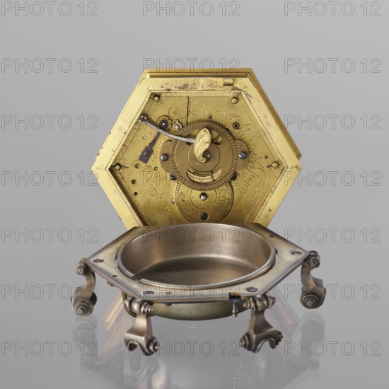 Tiled clock by Michael Gugenmus 1705–1775, maker/watchmaker. Rococo style with Regency ornament, 2nd half of the 18th century. Hexagonal table clock of gilded bronze on six volute legs, glazed side walls; silver-plated ring-shaped dial with black Roman numerals for hours and Arabic minutes, two winding holes. Center engraved Regency-Rococo cartouche with the signature Gugenmos/Varssavie. Hinged flap closing the mechanism; on the front plate of the mechanism a modest bun and a hammer with an engraved bronze head. Signature: Johann / Michael / Gugenmus / A Varsovie. Mechanism with strike repeater and a handle on the side wall under the upper edge. Timepiece; height 8.5 cm, width 13.5 cm, depth 13.5 cm. Place of origin: Warsaw, Poland., A hexagonal metallic object with a visible inner mechanism in gold tones is positioned upright behind a round shallow bowl-like receptacle that rests on an ornate stand; the stand and bowl show metallic colors including silver, bronze, and brass with areas...