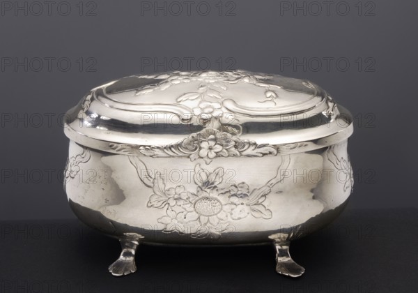 Sugar bowl by Hermann II Neupert fl. 1762–1775, rocaille ornament, rococo style; egg-shaped sugar bowl on four legs shaped as stylized lion's paws; belly and cover decorated with semi-plastic bouquets of flowers and rocailles; dated 1763–1775, Berlin, Germany; silver vessel, height 8.6 cm, width 13.5 cm, depth 8.7 cm, weight 255 g., A polished metal box with a domed lid sits on a dark surface against a dark gray background; the box has an ornate floral relief design on the lid and body, curved decorative edges, and stands on a group of small sculpted feet. The visible colors are silver, dark gray, and black.