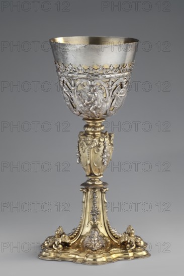 Mass chalice by Schwestermüller, David I fl. 1628–1678, goldsmith. Ovoid nodus and goblet with an openwork basket carved with Passion scenes: Prayer in the Garden of Olives, Ecce Homo, Christ in the Mystical Winepress, and angels bearing the Arma Christi. The profiled multi-leafed plinth and quadrangular foot are decorated with relief winged angel heads in conch-volute reserves, with hanging campanulas and applied angel heads on the division edges. Auricular ornamentation shows Netherlandish sources; figural depictions reflect features of late 16th-century Italian engravings. Vasa sacra liturgical vessel, height 25.5 cm, width 15.1 cm; ca. 1630–1635, Augsburg, Germany., A tall ornate goblet with a metallic bowl and a decorative stem and base; the bowl is primarily silver in color with intricate openwork and sculpted figures around its lower portion, and the interior rim shows a muted gold tone; the stem and base are predominantly gold in color with elaborate relief decorations, small s...