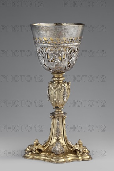 Mass chalice by David I Schwestermüller fl. 1628–1678, goldsmith. Liturgical vessel vasa sacra, ca. 1630–1635. Height 25.5 cm, width 15.1 cm. Mass chalice on a profiled multi-leafed plinth and a quadrangular foot decorated with relief winged angel heads in conch-volute reserves, above which hang campanulas and plastic angel heads set on the edges of the divisions; ovoid nodus with similar decoration. A goblet with an openwork basket with carved Passion themes — the Prayer in the Garden of Olives Agony in the Garden, Ecce Homo, Christ in the Mystical Winepress — among which are figures of angels with the Arma Christi. Chalice depicts Christ as the man of sorrows with instruments of the Passion. Graphic patterns likely compile several engravings; auricular ornamentation shows Netherlandish sources, while the depictions show features of late 16th-century Italian engravings cf. Agostino Carracci's Ecce Homo, 1587, after Corraggio. Place of origin: Germany, Augsburg., A tall ornate metal go...