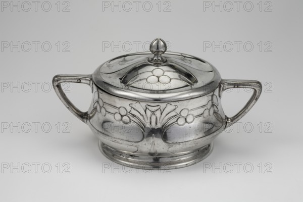 Art Nouveau sugar bowl by Berndorfer Metallwarenfabrik Arthur Krupp Berndorf; silver and metalworks; fl. 1843-, Art Nouveau style. Plant ornament decoration: circular sugar vessel on a low base with a body tapering toward the top, decorated with stylized leafy branches with circular fruit. Two handles decorated with profiles. Slightly arched lid with a bud-shaped handle. Circa 1900. Austria, Berndorf. Vessel. Height 10.2 cm; width 18.2 cm; depth 11.7 cm., A polished metal lidded container with a rounded body and a low pedestal base, shown against a pale gray background; the container has a domed lid topped with a small, rounded finial and a decorative raised motif on the lid, and a pair of angular handles extending horizontally from each side; the body features engraved floral and berry-like decorative patterns and smooth recessed oval panels, with visible wear marks and slight tarnishing on the reflective silver surface, showing shades of silver, gray, and hints of darker gray in the ...