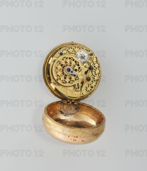 Pocket watch in case; maker: M. Cradelle fl. ca. 1750; watchmaker. Mythological scenes; rococo style. Watch in double case, outer case covered with lizard, with riveted rosette. Mid-18th century, London, England, Great Britain. Timepiece. Height 6 cm; width 4.5 cm; dial diameter 3.5 cm., A small open circular metal object with an ornate upper interior showing intricate filigree and gear-like components in bright gold tones with small dark accents and a central jewel-like element in deep red, attached by a hinge to a rounded lower bowl-shaped cavity that is also metallic gold with areas of darker tarnish and a small patch of reddish markings on its outer side; the background is a pale neutral gray.