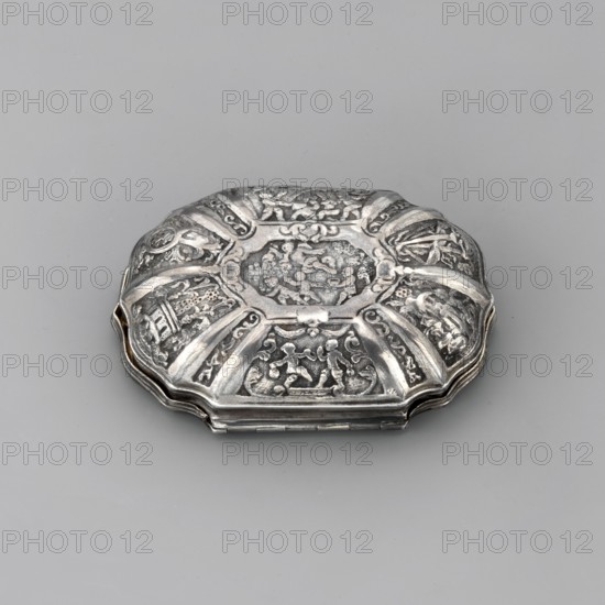 Snuffbox, mythological scenes: Venus, Cupid, Mars, putti. Oval snuffbox with irregular edges; on the lid within a smooth border a scene depicting a seated man and a semi-nude woman glancing into a mirror while Cupid stands before them handing the man a swaddled child—possibly alluding to the romance of Venus and Mars. Around, in rhomboidal reserves, scenes of fighting putti, Regency ornaments and intertwined dolphins; on the bottom reserve playing putti, above them a duel scene, below two knights in a galley; on the sides ships and decorations of Regency ornaments. Gilded interior. 1720s–1730s, France. Smoking accessory. Height 6.5 cm; width 5 cm; weight 49.5 g., A decorative metal box with an octagonal, scalloped shape rests on a smooth light gray surface; the box has a raised ornate lid divided into radiating panels filled with intricate embossed scenes and motifs, giving varied textures of carved relief and smooth bands. The metal shows a silvery color with darker gray areas in the ...