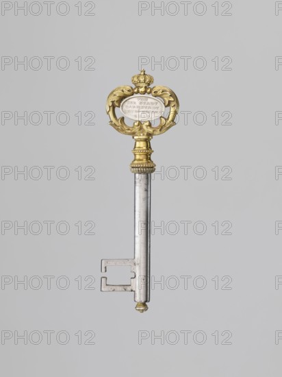 Souvenir key, steel, with a full shank, with a simple feather in the form of two square brackets. Gilded bronze knot and handle: oval handle, composed of two U-shaped sections decorated with leaves, fastened with a ducal mitre. Inside, an oval plaque on one side with the Gothic monogram L and red enamel, on the other with a cast inscription: VON / DER STADT / DARMSTADT / DEN 26 DECEMB: / 1833. Given to Duchess Mathilde Caroline of Bavaria 1813–1862 and her husband Ludwig III 1806–1877 on the occasion of their wedding by the city of Darmstadt to the donated house in Mathildenhöhe, 1833, Germany. Varia. Height 8.9 cm, width 2.6 cm., The image shows a single ornate key centered on a plain light gray background; the key has a metallic shaft in a silver-gray color and an ornate bow and collar in a gold color with decorative scrollwork and a crown-like motif, and there is a small rounded gold-colored finial at the bottom of the shaft.