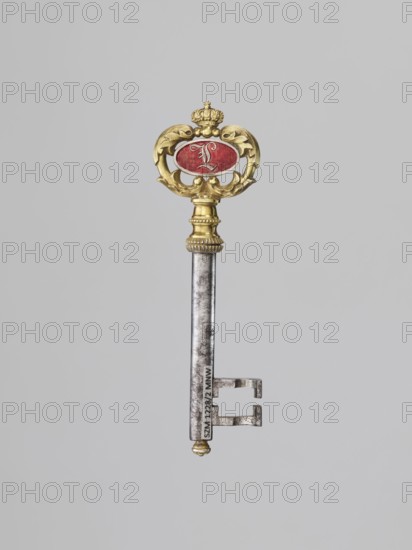 Souvenir key, steel, with a full shank and a simple feather in the form of two square brackets. Gilded bronze knot and handle: oval handle composed of two U-shaped sections decorated with leaves, fastened with a ducal mitre. Inside, an oval plaque on one side with a Gothic monogram L in red enamel, on the other with a cast inscription: VON / DER STADT / DARMSTADT / DEN 26 DECEMB: / 1833. Given to Duchess Mathilde Caroline of Bavaria 1813–1862 and Ludwig III Grand Duke of Hesse, 1806–1877 on the occasion of their wedding, 26 December 1833; Germany. Varia; height 8.9 cm, width 2.6 cm., A single ornate key is shown against a plain light gray background; the key has a metallic silver shaft and bit with visible surface texture and a decorative gold-toned bow and collar, and the bow surrounds an oval red element.