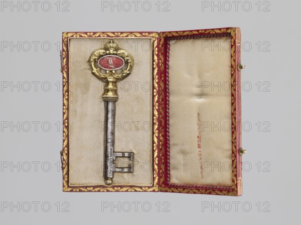 Souvenir key, steel, full shank with a simple feather in the form of two square brackets. Gilded bronze knot and handle: oval handle composed of two U-shaped sections decorated with leaves, fastened with a ducal mitre. Inside, an oval plaque: on one side a Gothic monogram L in red enamel; on the other a cast inscription: VON / DER STADT / DARMSTADT / DEN 26 DECEMB: / 1833. Given to Duchess Mathilde Caroline of Bavaria 1813–1862 and her husband Ludwig III Grand Duke of Hesse; 1806–1877 on the occasion of their wedding by the city of Darmstadt, 1833. Germany. Varia. Height 8.9 cm; width 2.6 cm., A decorative metal key rests in an open rectangular box lined with pale beige fabric, the key featuring a golden ornate bow with a central oval panel in red and a long silver shaft; the box interior edges are bordered with red trim that has gold decorative patterns, and the right side of the box shows folded and slightly wrinkled beige lining with red trim and gold accents; the surrounding backgr...