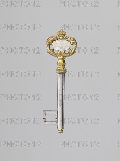 Souvenir key, steel, with a full shank and a simple feather in the form of two square brackets. Gilded bronze knot and handle: oval handle composed of two U-shaped sections decorated with leaves, fastened with a ducal mitre. Inside, an oval plaque with a Gothic monogram M and blue enamel on one side, and a cast inscription on the other: VON / DER STADT / DARMSTADT / DEN 26 DECEMB: / 1833. Given to Duchess Mathilde Caroline of Bavaria 1813–1862 and her husband Ludwig III on the occasion of their wedding, 1833. Germany; Darmstadt. Varia. Height 8.9 cm, width 2.6 cm., A single ornate key is pictured against a plain light gray background; the key has a long metallic silver shaft with a rectangular bit at the lower end and a small rounded tip with a touch of gold, and the upper section features a decorative gold head with curved foliate shapes and a small crown-like element, with accents of white visible within the central area of the head.